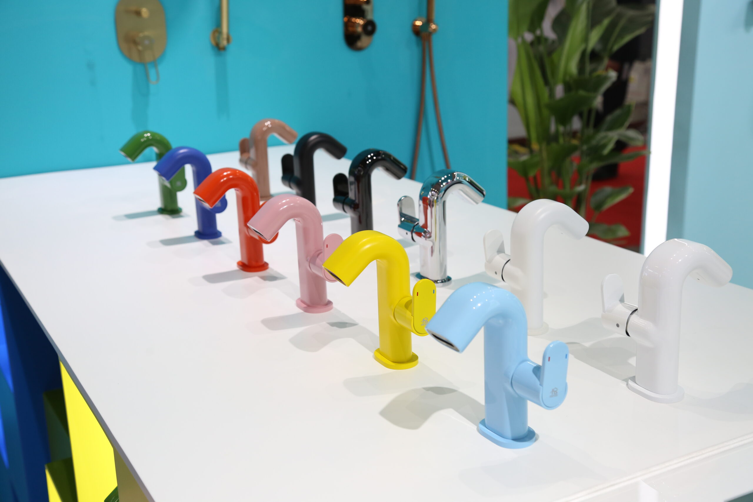 Multicolor designer faucets on white counter: Emerald Green, Coral Pink, Chrome, Matte Black. Pantone-certified finishes with anti-fingerprint nano-coating. Arranged in chromatic sequence against teal feature wall.